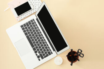 Un ordinateur portable ouvert vu de dessus, avec un smartphone posé sur un carnet à motifs, une tasse contenant des ciseaux et des stylos, et une petite bougie blanche sur un bureau beige.