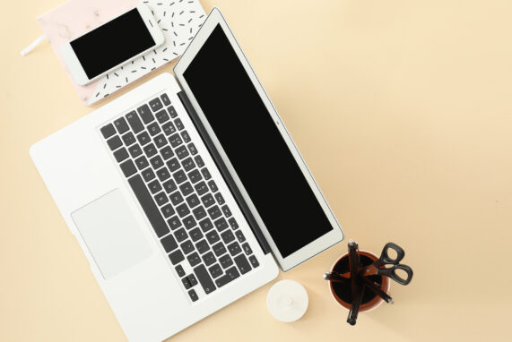 Un ordinateur portable ouvert vu de dessus, avec un smartphone posé sur un carnet à motifs, une tasse contenant des ciseaux et des stylos, et une petite bougie blanche sur un bureau beige.