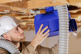 Un ouvrier portant un casque de protection blanc installe un système de ventilation bleue sous un plafond en bois, avec des conduits flexibles reliés à l’appareil.