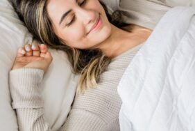 Femme souriante allongée dans un lit, les yeux fermés, semblant profiter d’un sommeil paisible sous une couverture blanche.